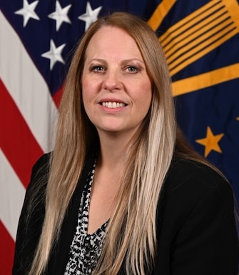 Portrait of person in professional attire with long hair in front of two flags.