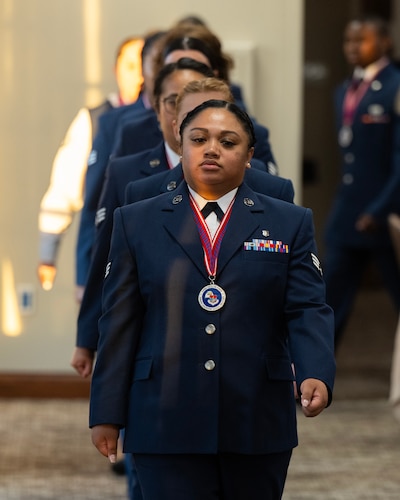 A woman in dress uniform, and wearing a medallion, lead a line of Airmen marching into a room.