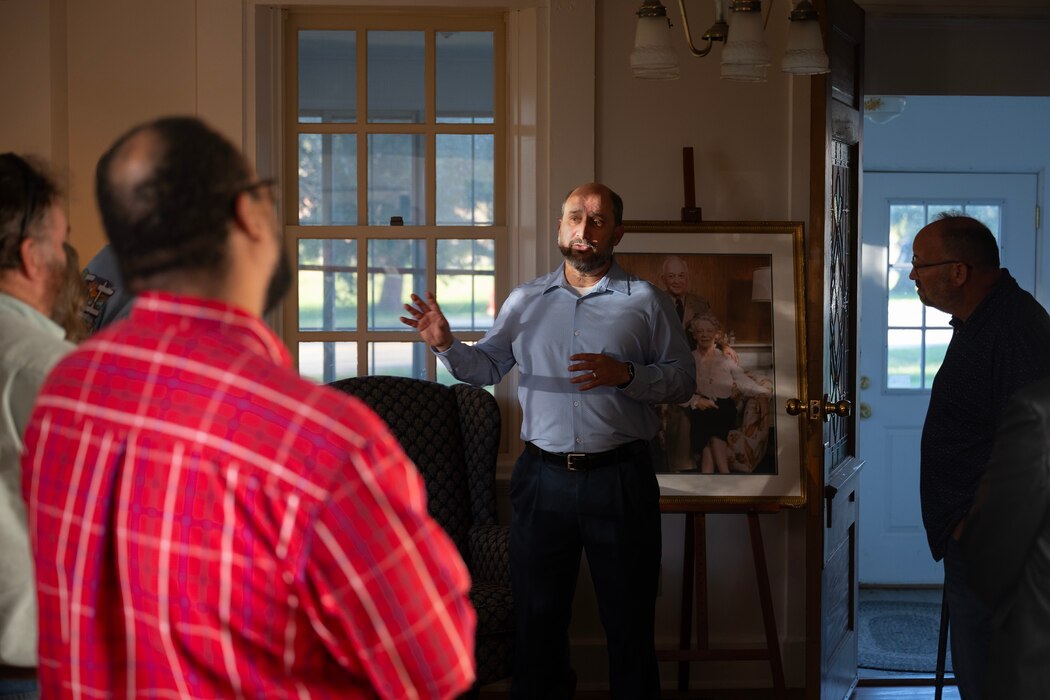 A bearded man, in a blue shirt, stands in an old-style living room talk to people gathered around him.