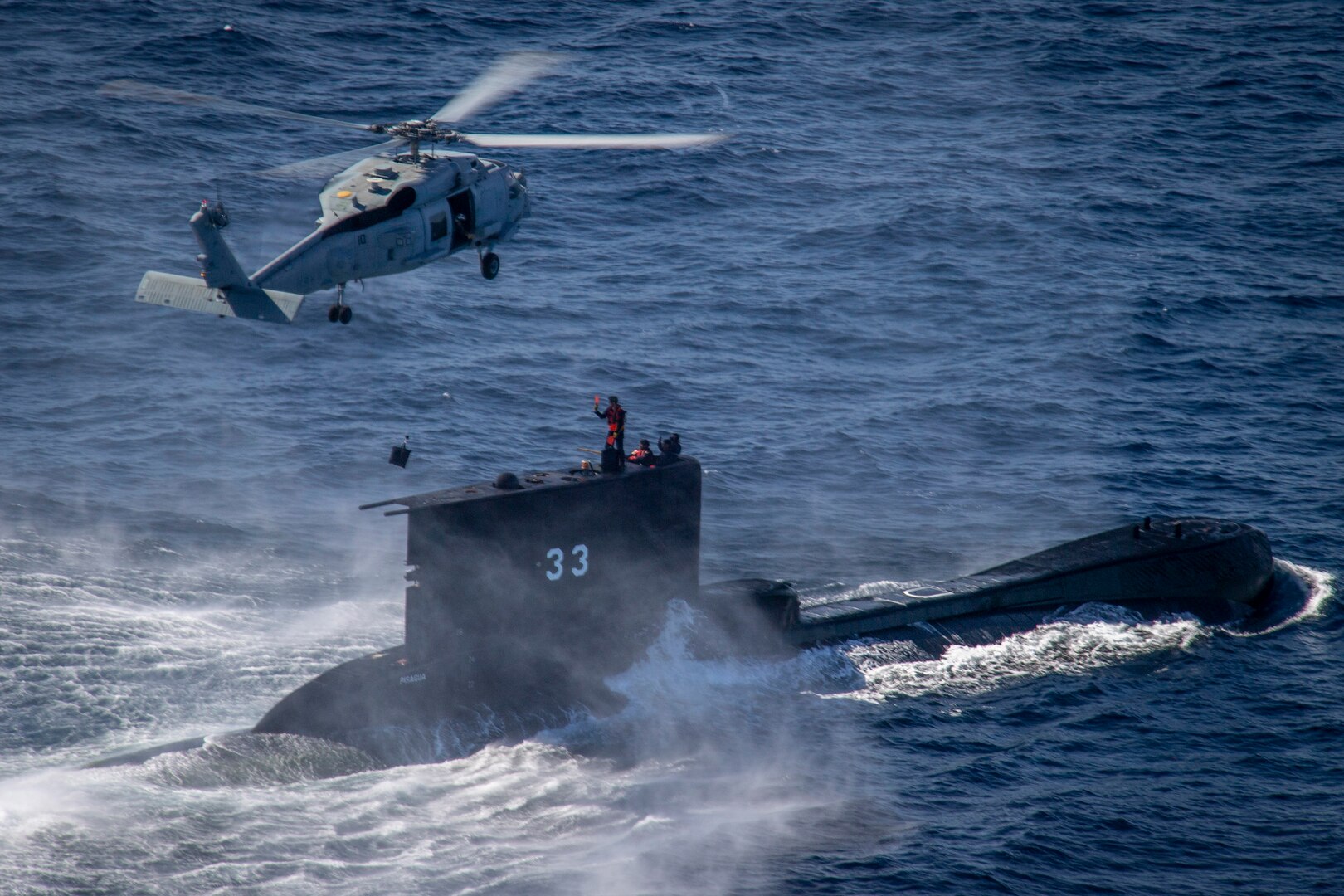 A helicopter hovers over a service member standing on a submarine in a body of water during daylight.