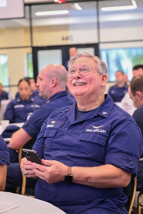 A group of individuals seated at tables all in the background while Wilson Riggan a Coast Guard Auxiliarist is pictured looking up smiling at the stage.