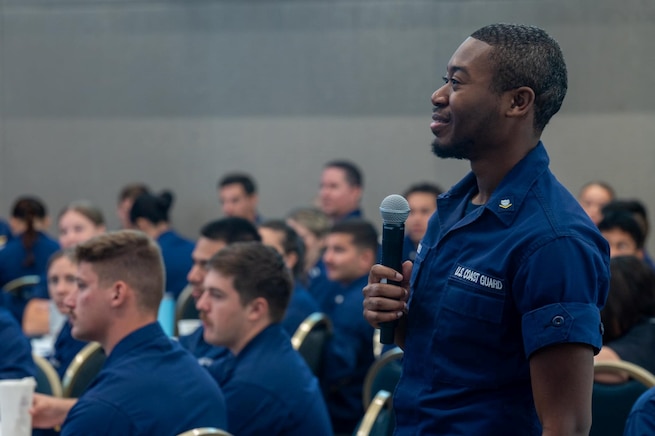 A group of Coast Guardsmen seated at tables, with one individual standing up in the crowd with a microphone in the process of asking a question.