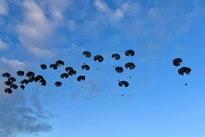 Multiple black parachutes carrying containers of supplies drop to the ground against a blue sky.