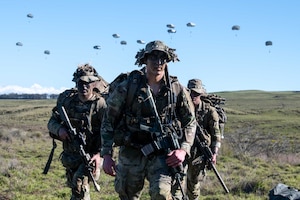 Three soldiers carrying weapons walk toward the camera in a large field while parachutes drop from a blue sky behind them.