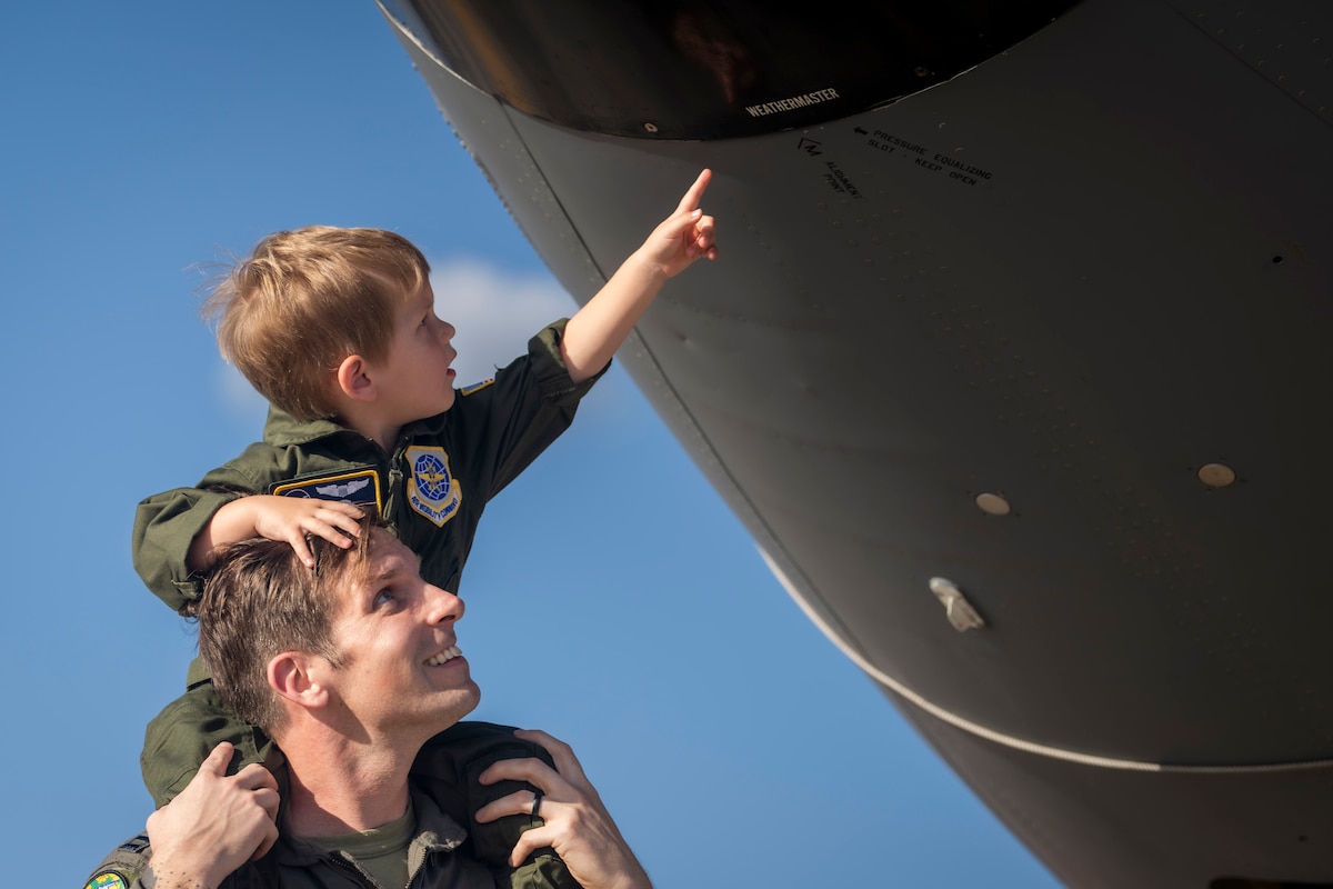 A child sits on the shoulders of a pilot and points to a part on an aircraft.