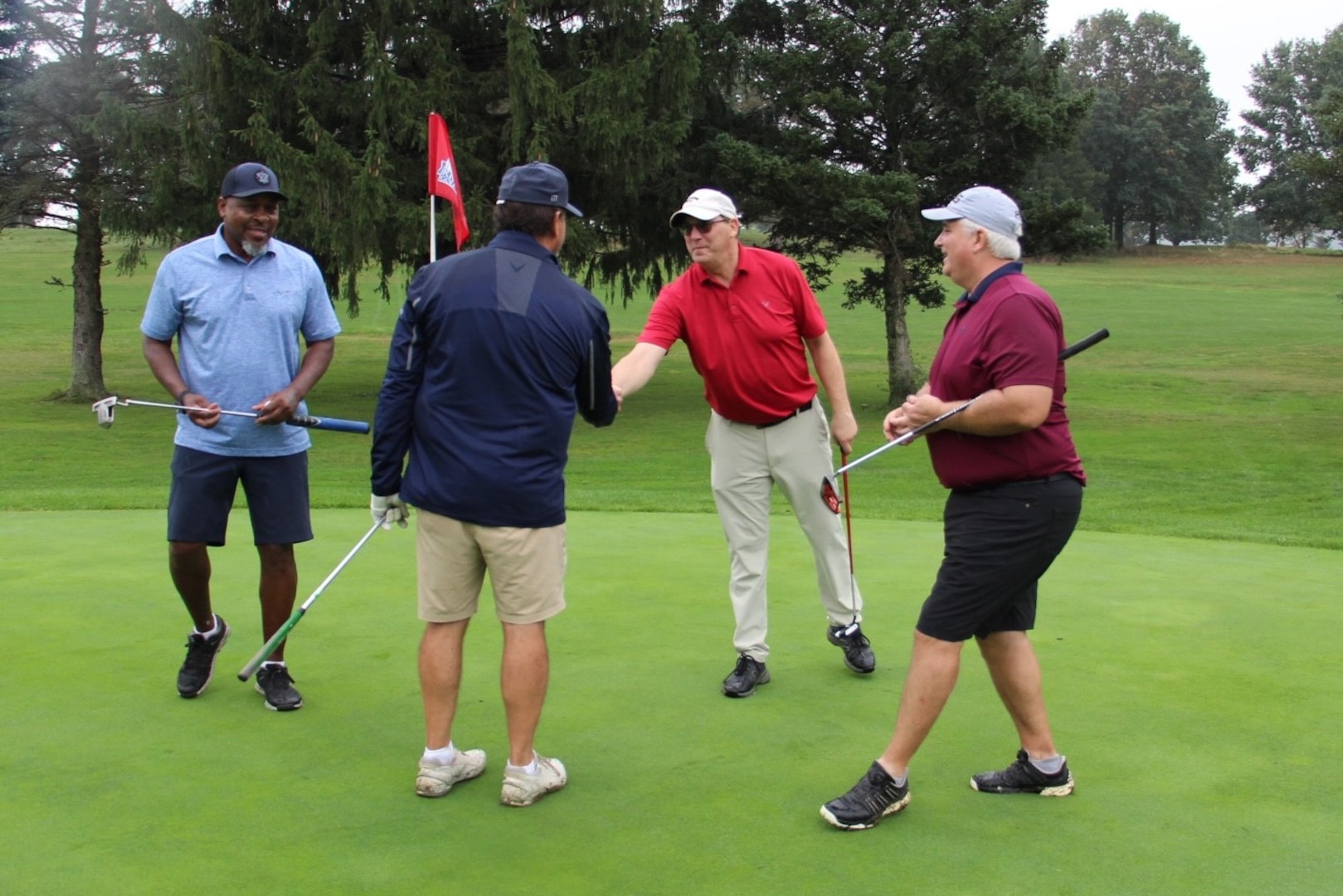 Four golfers gather around a hole on a golf course. The two in the middle are shaking hands.