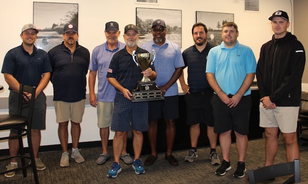 Eight men pose for a group photo with the man in the middle holding a large trophy.