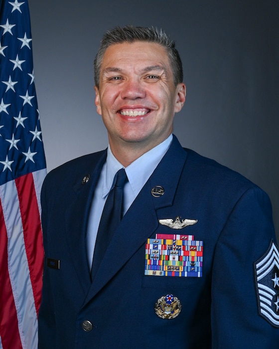 A man in a Air Force service dress poses in front of an American flag and a gray background.