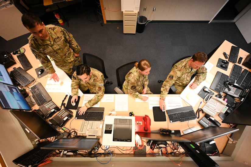 A group of airmen and guardians sit at a long desk looking at computer monitors.