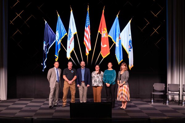 Civilians wearing business attire pose for a photograph on a stage.