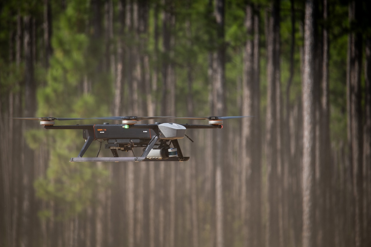 A drone flies level with a forest of green and brown trees in the background.