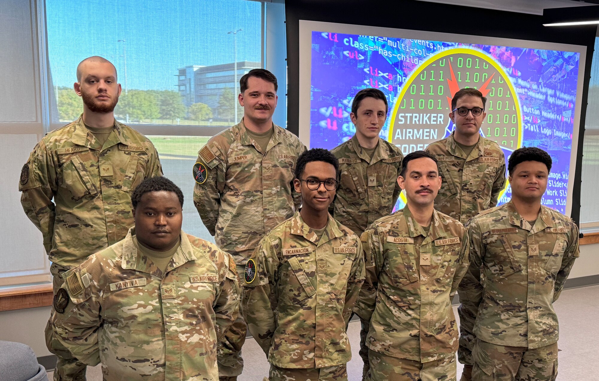People stand in a group of two rows to pose for a group photo. The projector screen behind them displays the banner and shield for AFGSC's Striker Airmen Coder Program.