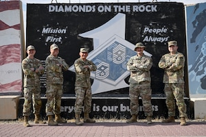 Five first sergeants stand in front of a first sergeant mural that features the three ranks commonly worn by first sergeants.