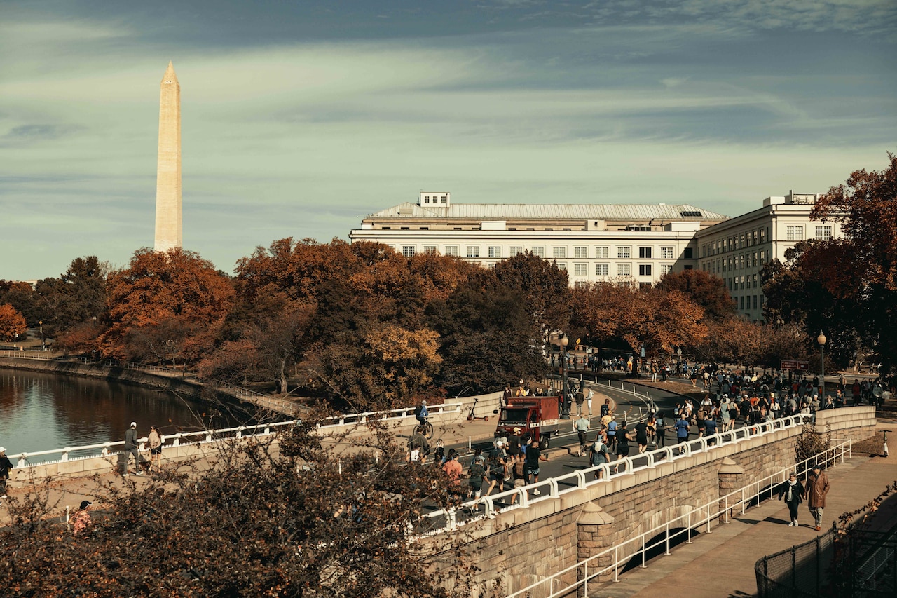 A large group of people run and walk along a city street with a monument in the background.