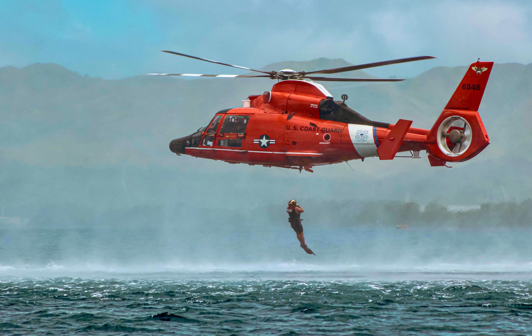 An MH-65 Dolphin helicopter crew trains over Apra Harbor, Guam, on March 14, 2023. The MH-65 Dolphin helicopter is a short-range recovery helicopter used by the crew to perform search and rescue, law enforcement, and homeland security missions. The aircrew is forward deployed to Guam from Hawaii for six weeks serving the people of Guam and the Commonwealth of the Northern Marianas Islands while working with U.S. Coast Guard Forces Micronesia/Sector Guam to assess the feasibility of more frequent operations in the islands and provide an additional search and rescue asset as available. (U.S. Coast Guard photo by Petty Officer 2nd Class Ed Mewborn)