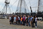 A group of DCMA employees stand before the wood-hulled USS Constitution in Boston Harbor.