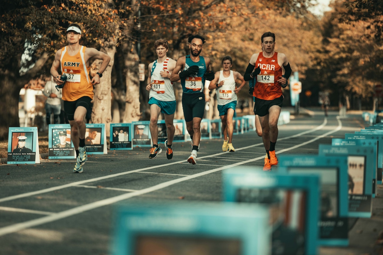 A group of athletes run along a street with large photographs along the side.