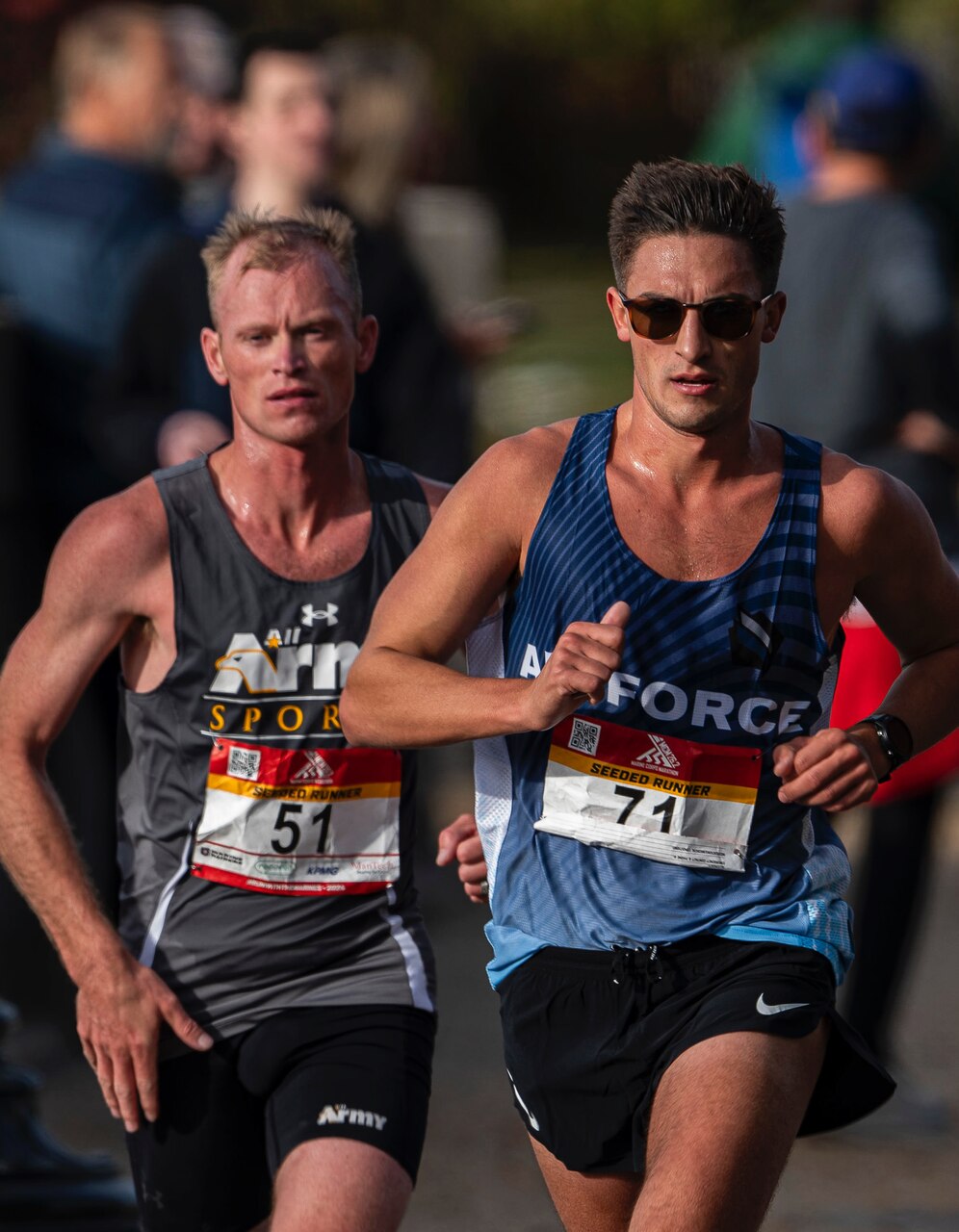 Two men run during a marathon race. A crowd of people are blurred in the background.