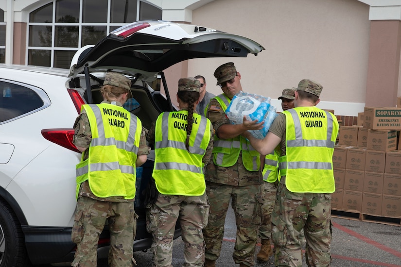 Several National Guardsmen wearing neon vests over their camo load a case of water and other items into the open back of an SUV.