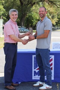 Two men stand outside holding a small trophy.