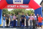 A group of people stand outside under an inflatable race start/finish line arch, the man in the center is holding a large trophy and wearing a camouflage military uniform.