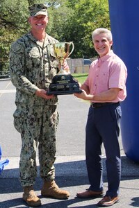 Two men, one wearing a camouflage uniform, stand outside holding a large trophy.