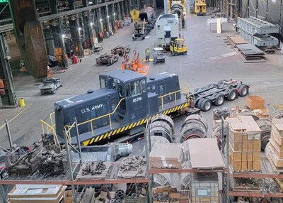 A view from above showing the inside of a large workspace. The Diesel locomotive sits in the center of the photo being unloaded from a semi truck trailer.