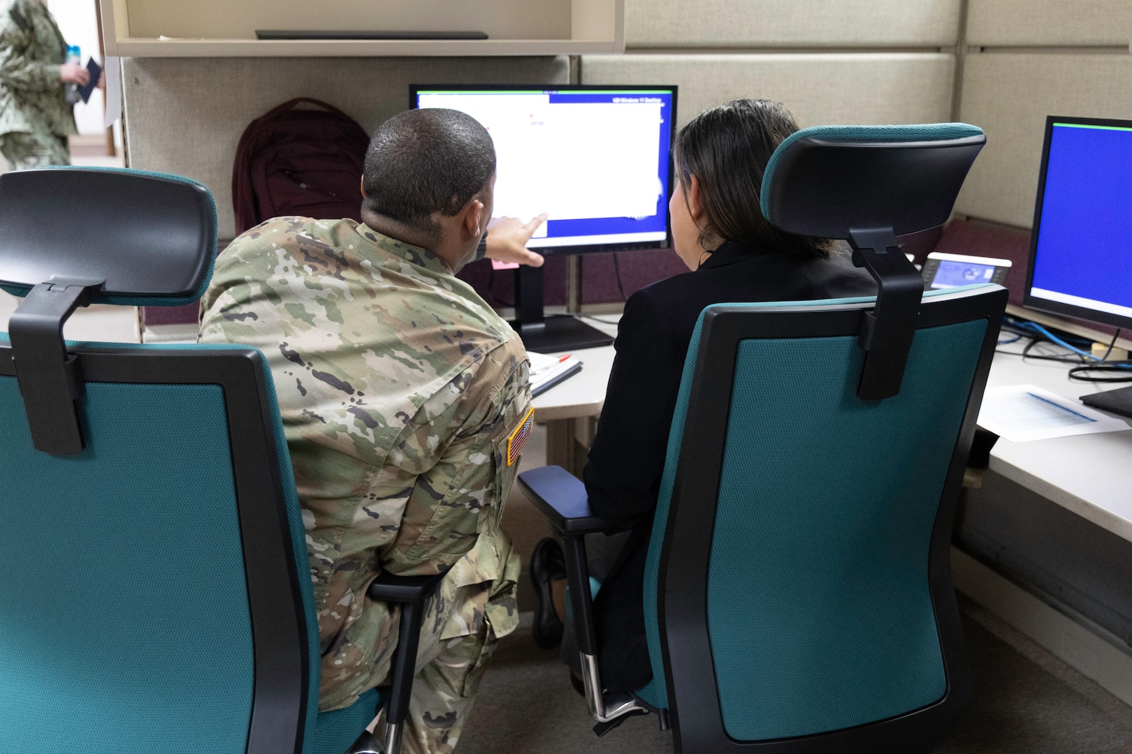 A dark skinned man points to a computer screen as he sits next to a light skinned dark haired woman in a cubicle. They are sitting in green chairs. He is in uniform and she is not.