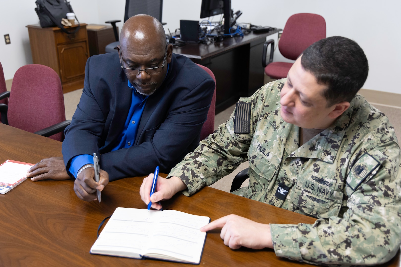 A dark skinned man in a dark blue business suit talks with a light skinned man with black hair in a green camouflage Navy uniform at a conference table. They are pointing to a book on the table.