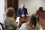 A light skinned man in a blue dress shirt and dark blue suit jacket with graying hair and beard talks with two women at a conference table. One has red curly hair and the other has blond and black straight hair. The curly haired woman has a light green jacket on and the other woman has a white sweater.