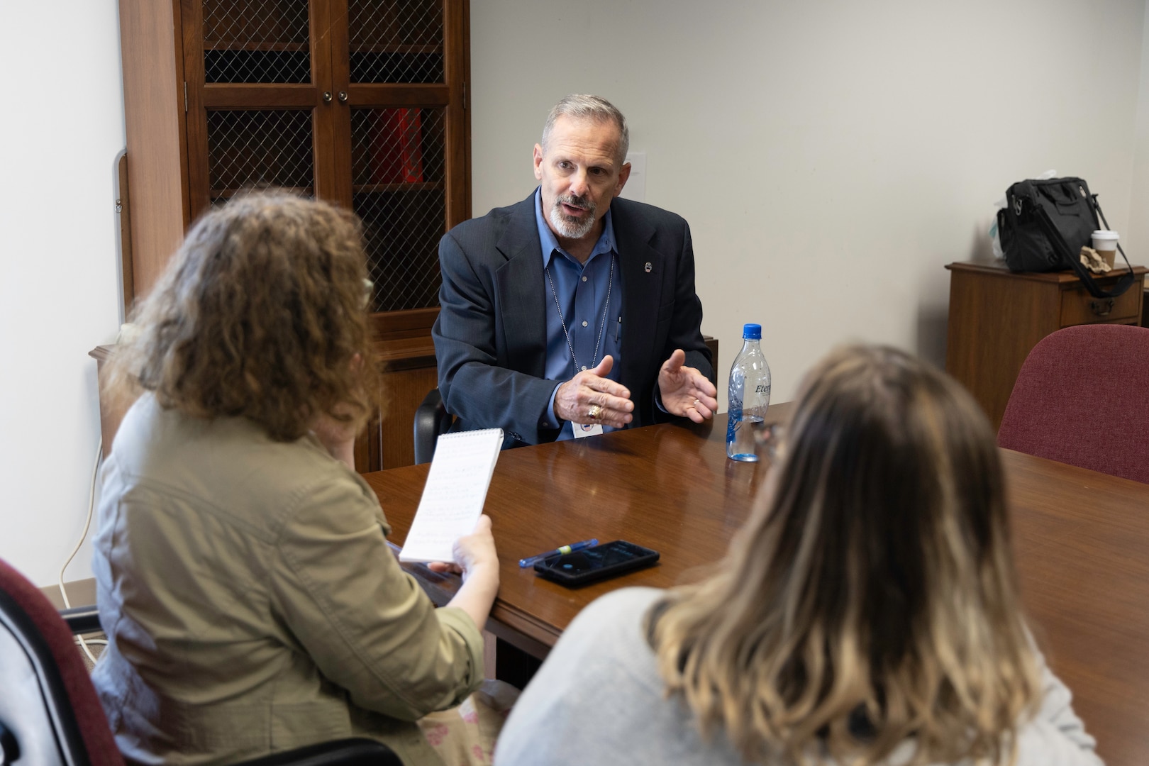 A light skinned man in a blue dress shirt and dark blue suit jacket with graying hair and beard talks with two women at a conference table. One has red curly hair and the other has blond and black straight hair. The curly haired woman has a light green jacket on and the other woman has a white sweater.