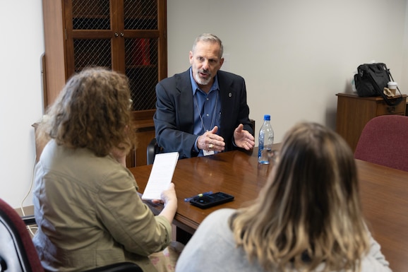 A light skinned man in a blue dress shirt and dark blue suit jacket with graying hair and beard talks with two women at a conference table. One has red curly hair and the other has blond and black straight hair. The curly haired woman has a light green jacket on and the other woman has a white sweater.