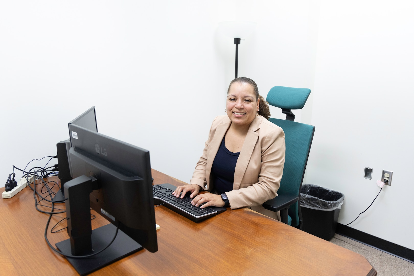 A dark skinned woman in a black dress shirt and tan suit jacket sits at a large brown desk in an office with white walls. She is smiling as she looks up from her computer monitor and has her hands on the keyboard.