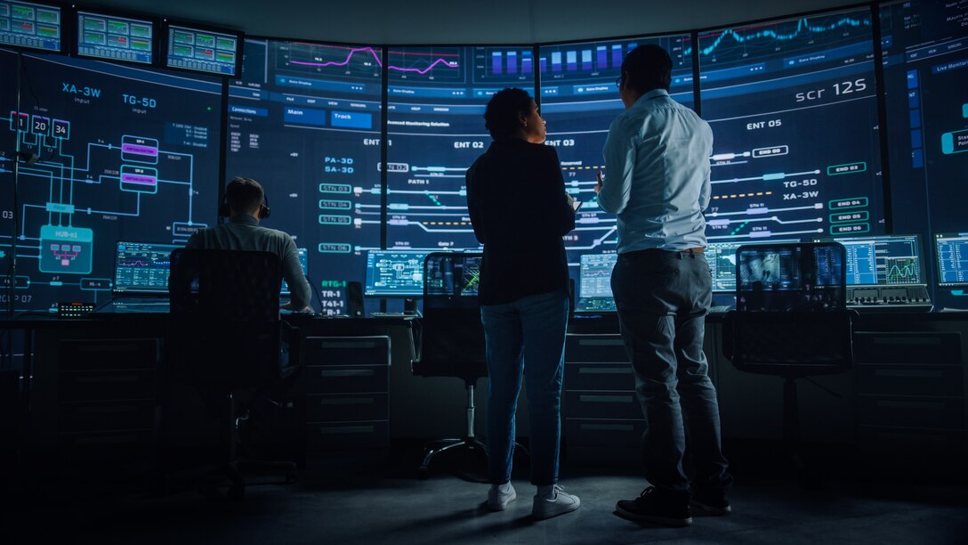A man and woman standing in front of a large screen displaying data on a screen, with a man wearing a headset working on a computer in the background