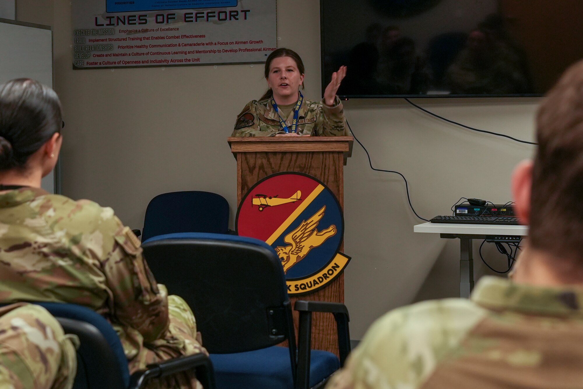 A woman in a military uniform stands at a podium to talk to a classroom of military students.