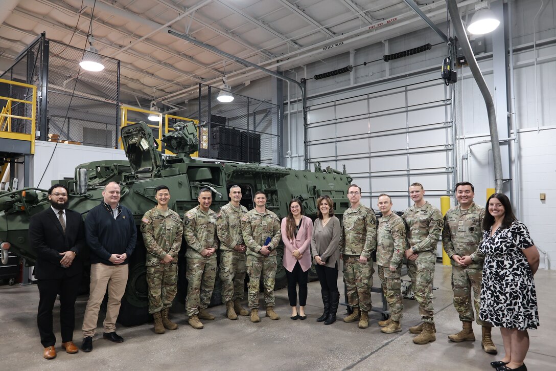 soldiers and civilians stand in front of an Army Stryker vehicle in a warehouse
