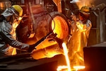 Two people in protective gear pour molten metal into molds.