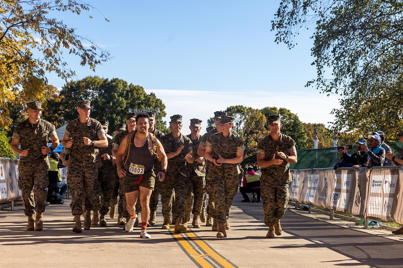 A marathon runner runs alongside a group of uniformed Marines on a road during a clear day.