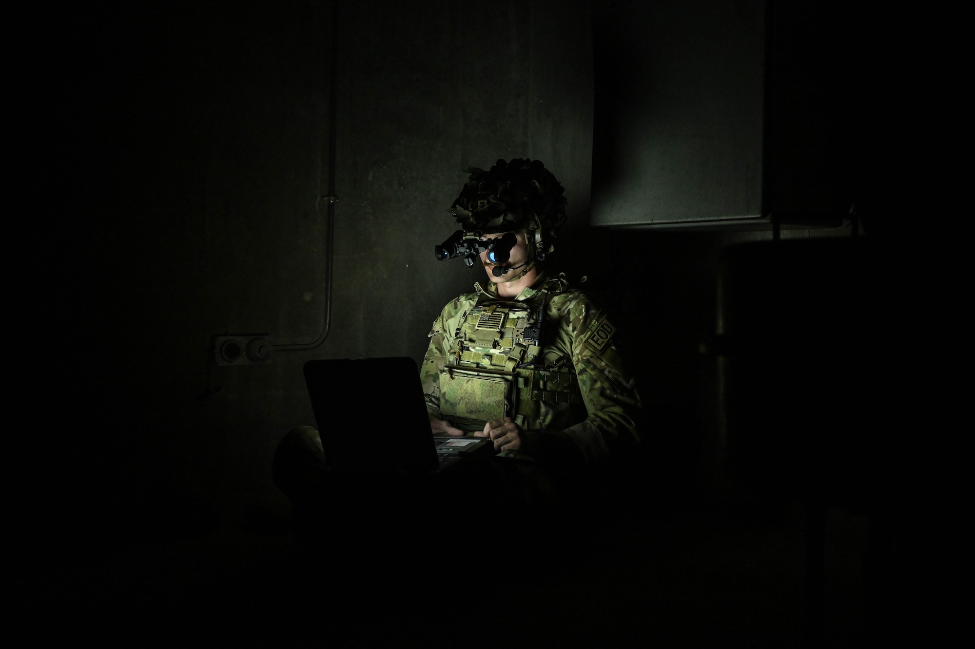 A man in uniform wearing night vision goggles looks down at his computer.
