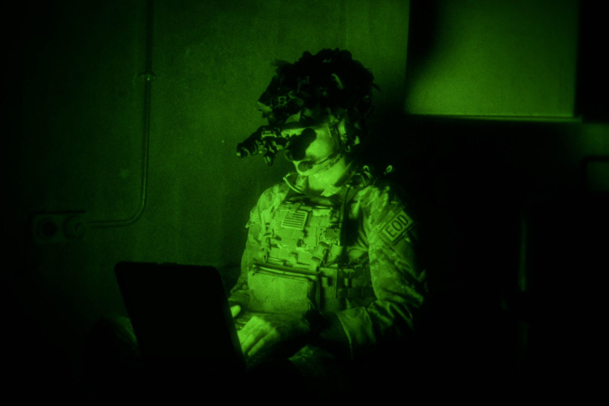 A man in uniform wearing night vision goggles looks down at a computer.