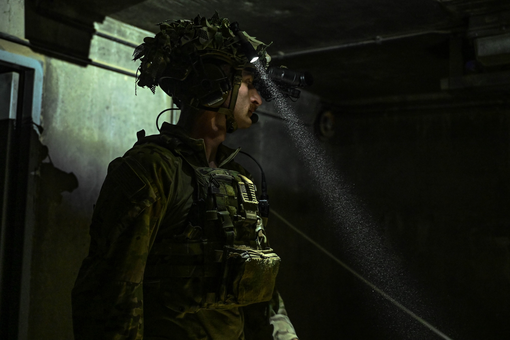 A man in uniform wearing a helmet mounted flashlight looks around a room.