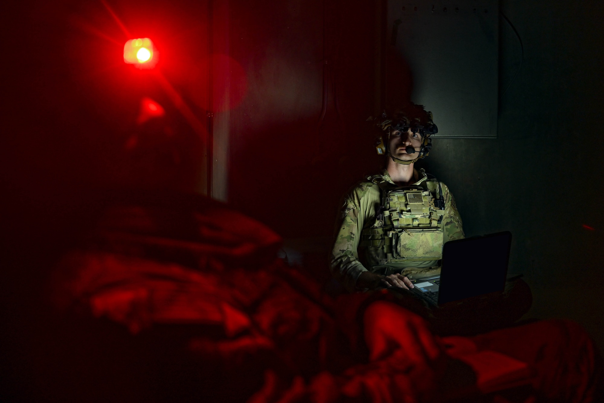 A man in uniform looks at another man in uniform while working on a computer in the dark.