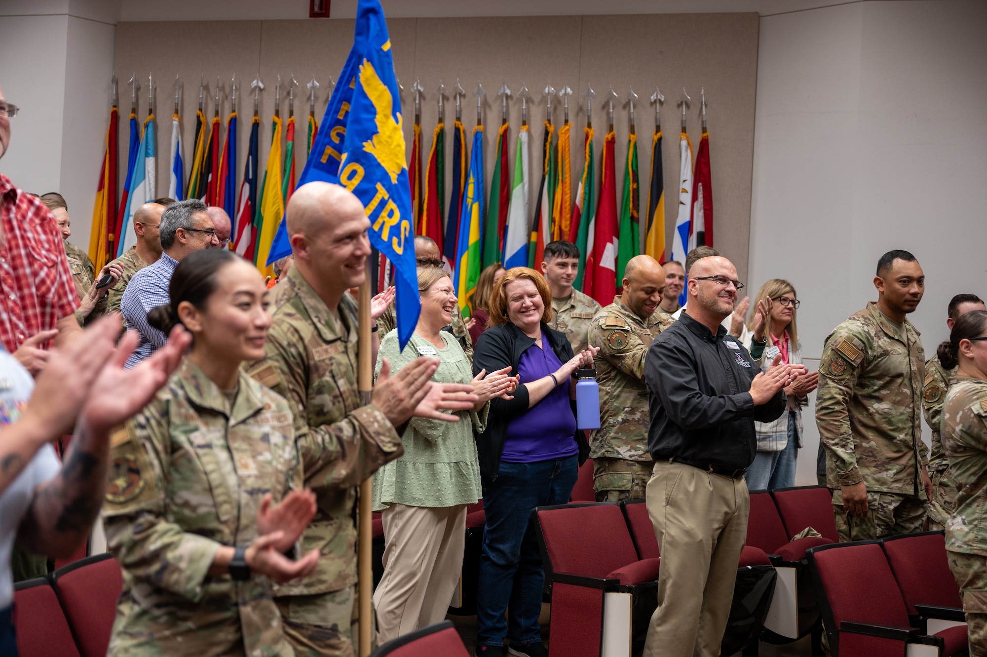 Uniformed members and civilians clap and laugh in an auditorium during an award ceremony.