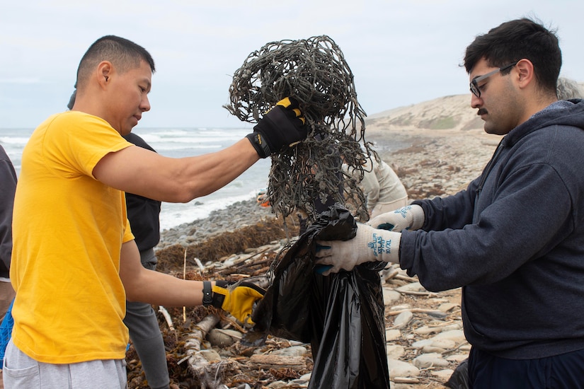 Two people put a dirty fishing net in a bag while standing on an island with the ocean behind them.