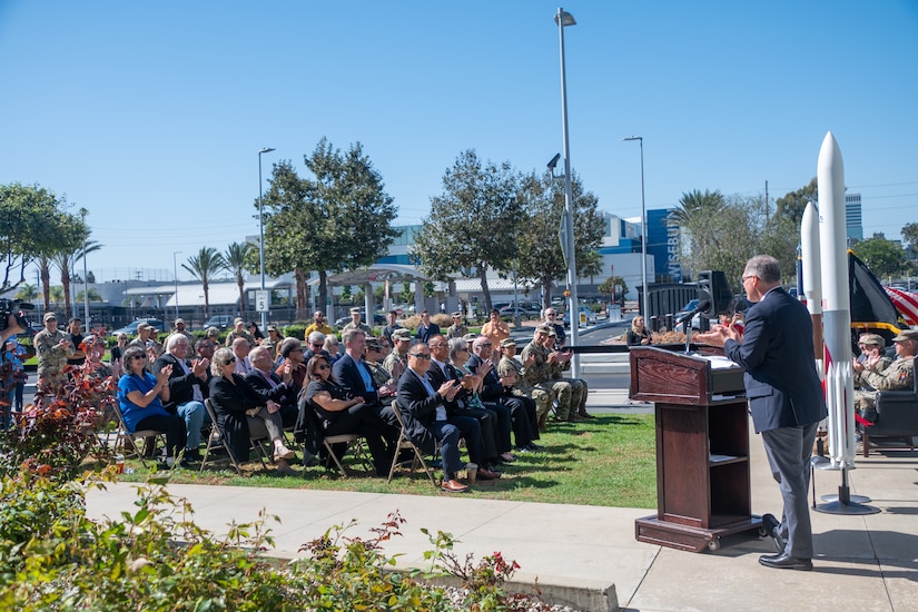 A group of people sit in chairs outside in front of small model rocket and a person wearing business attire speaking from a lecturn.