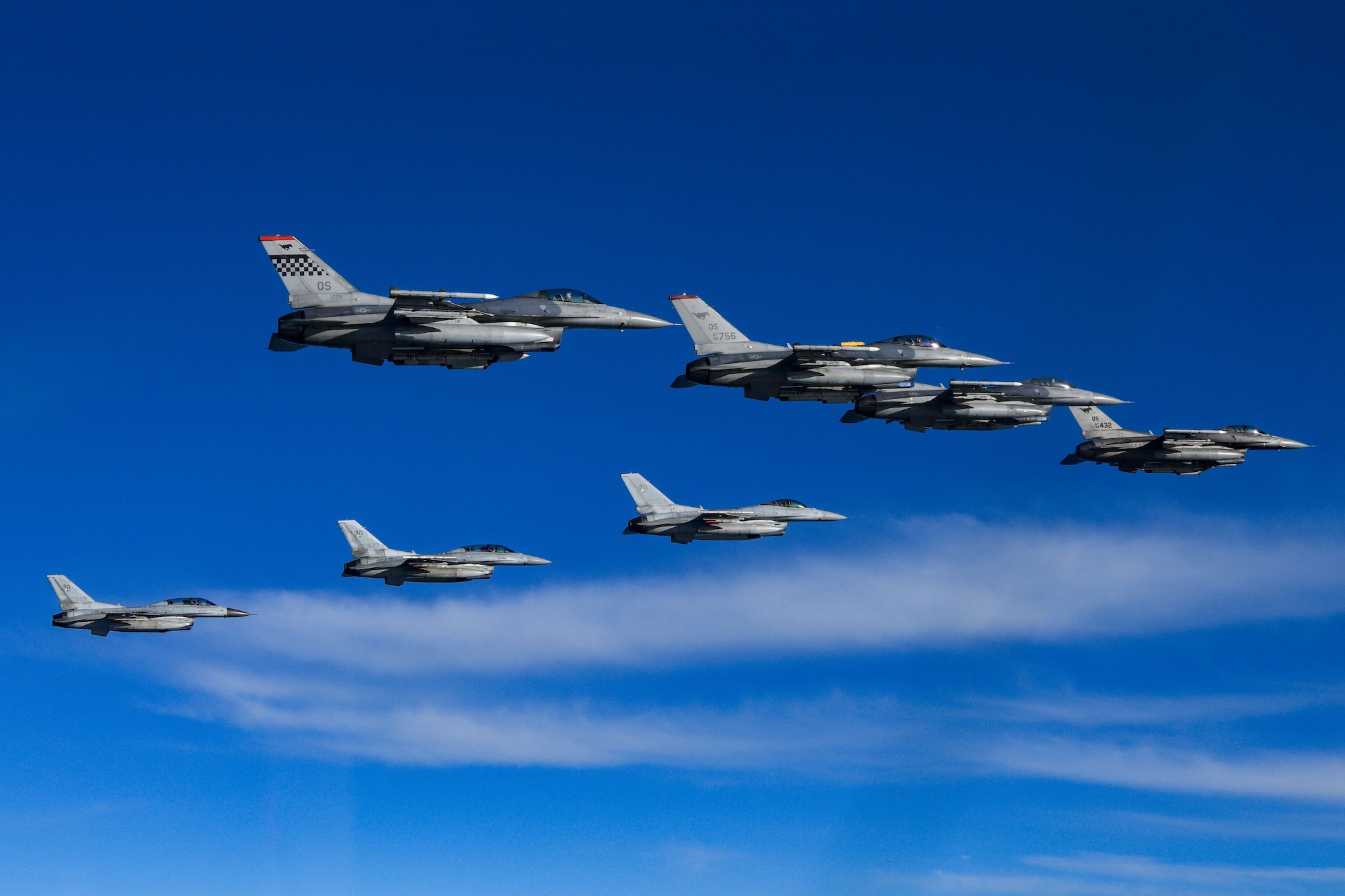 Eight F-16 fighter jets fly in a v formation over blue water with white clouds. The jets are seen from below and to the side.