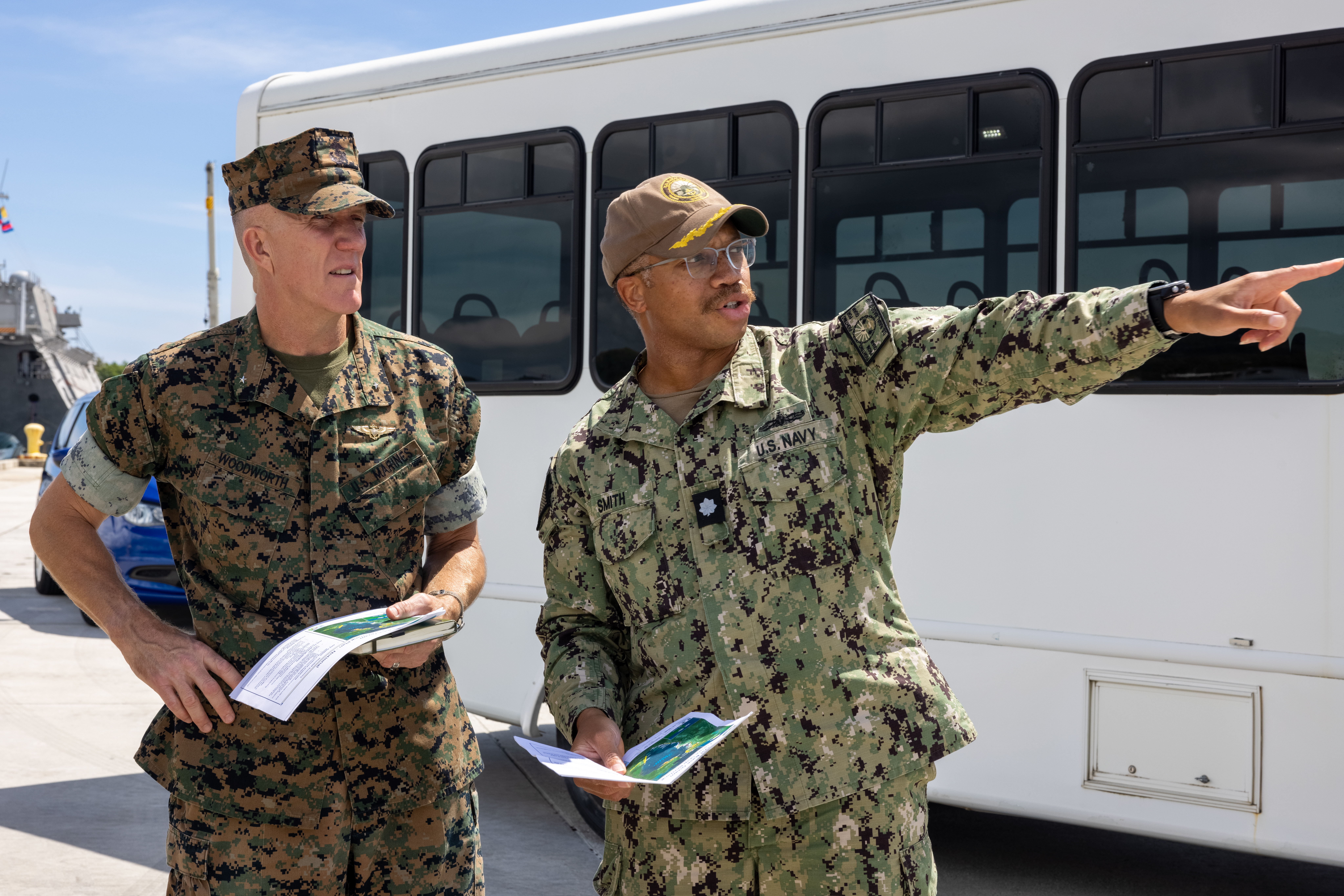Two Marines in front of a white bus, one at something to their right.