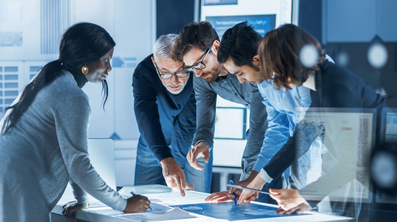 A group of five men and women, leaning over a table discussing and writing notes on a printed project design