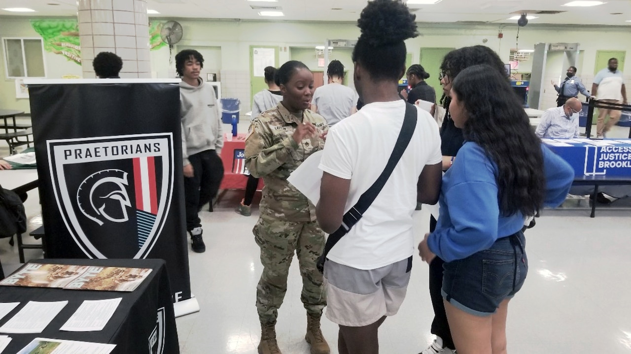 A soldier talks to three teenagers in a large room.