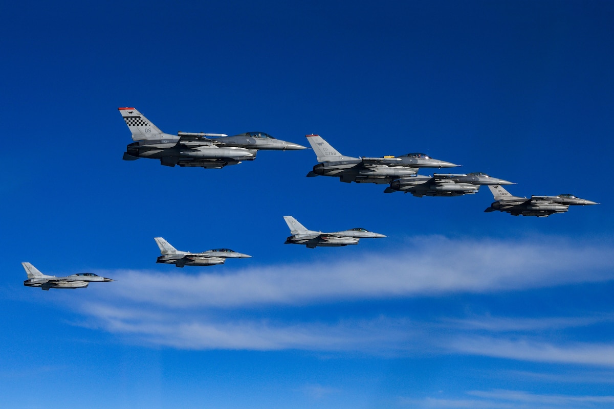 Eight F-16 fighter jets fly in a v formation over blue water with white clouds. The jets are seen from below and to the side.